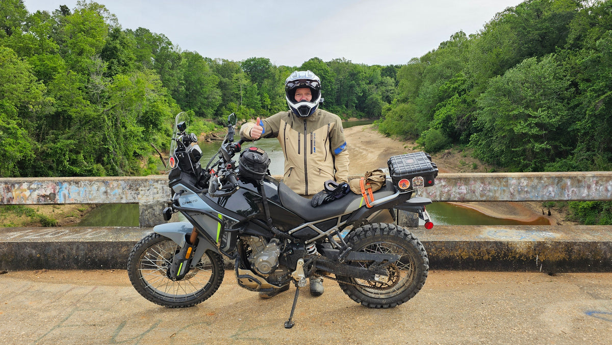 A person wearing a helmet and beige riding jacket stands beside a black and grayADV motorcycle on a bridge, with a river and green trees in the background.